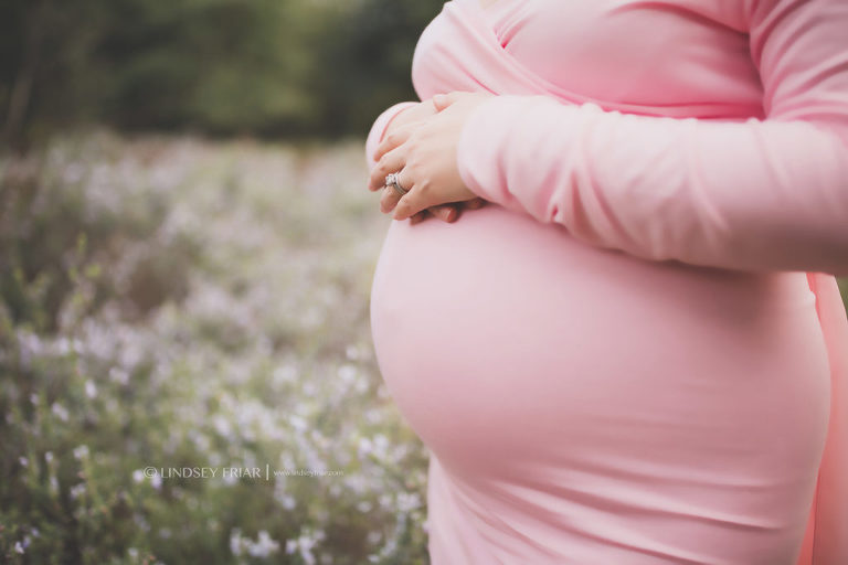 close up of a pregnant belly of a mama wearing pink standing in a field of flowers