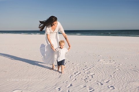 mom walking behind a baby holding his hands on the beach